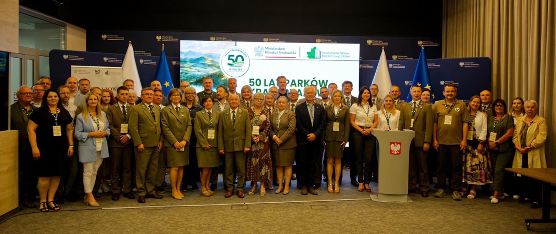 Grupowe zdjęcie uczestników obchodów 50-lecia parków krajobrazowych w Polsce, wykonane w sali konferencyjnej przed jubileuszową ścianką i flagami Polski oraz Unii Europejskiej.