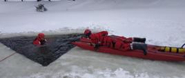 Zdjęcie przedstawia strażaków ubranych w suche skafandry. Na zdjęciu znajduje się dwóch strażaków. Jeden z nich znajduję się w wodzie, a drugi zbliża się do niego na saniach lodowych. W tle akwen pokryty lodem.