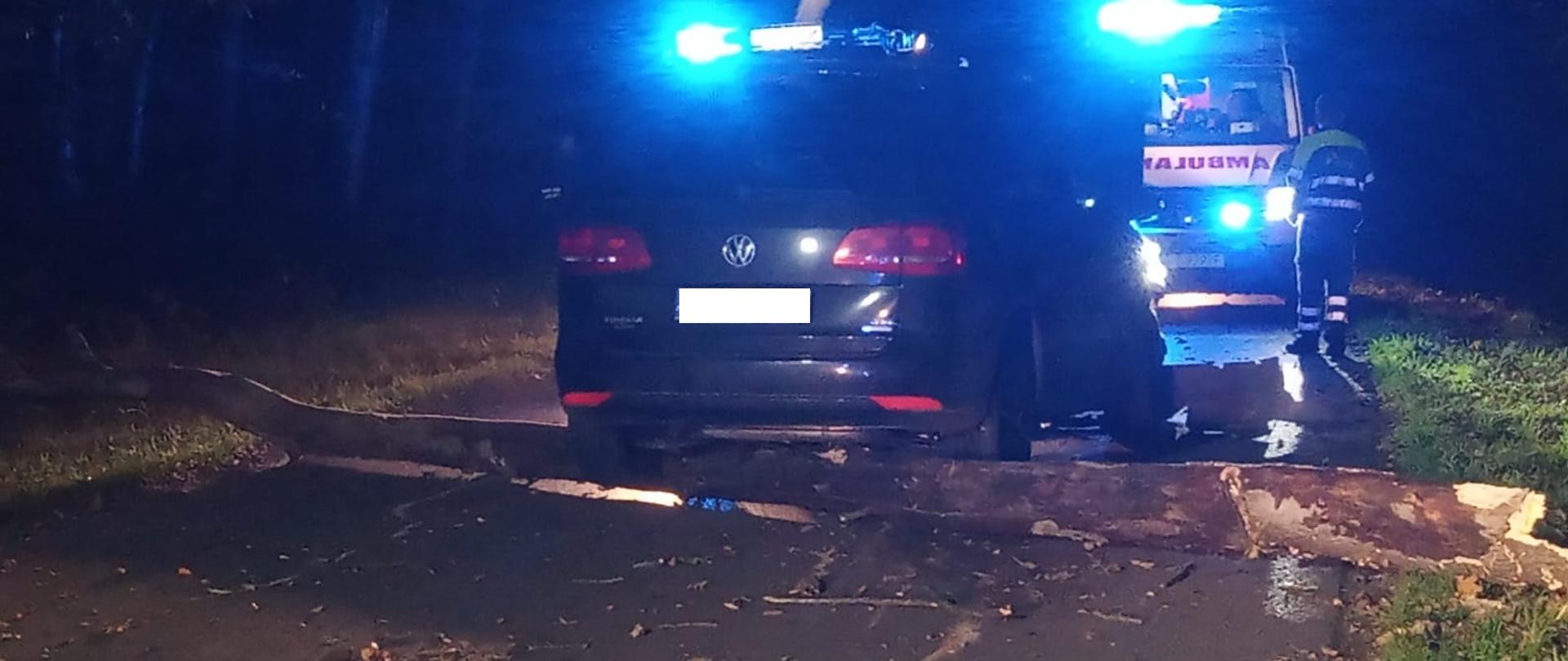samochód stoi środku drogi a drzewo leży pod pojazdem w oddali widać Karetkę Pogotowia Ratunkowego, wokół las.