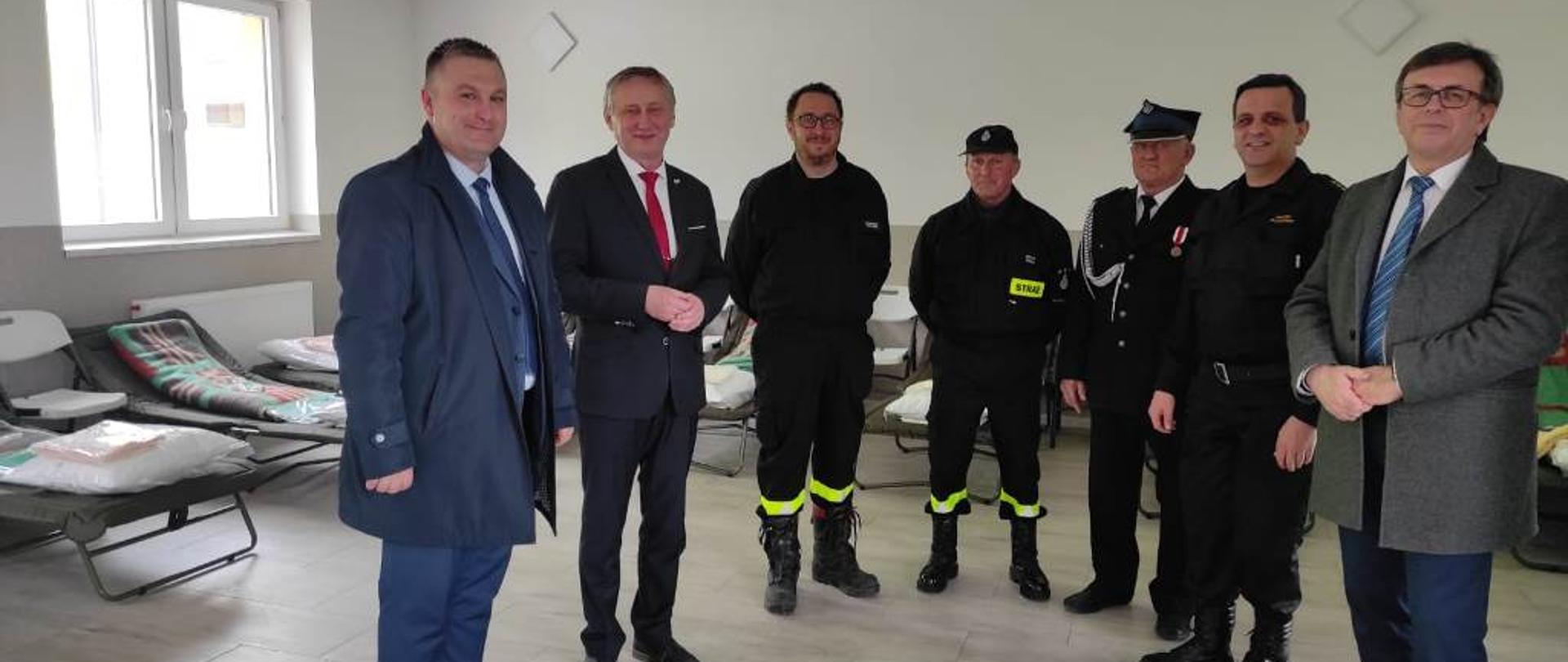 Przedstawiciel władzy rządowej oraz samorządowej w towarzystwie komendanta powiatowego i druhów strażaków w trakcie dokonywania przeglądu pomieszczeń i wyposażenia przygotowanego dla uchodźców