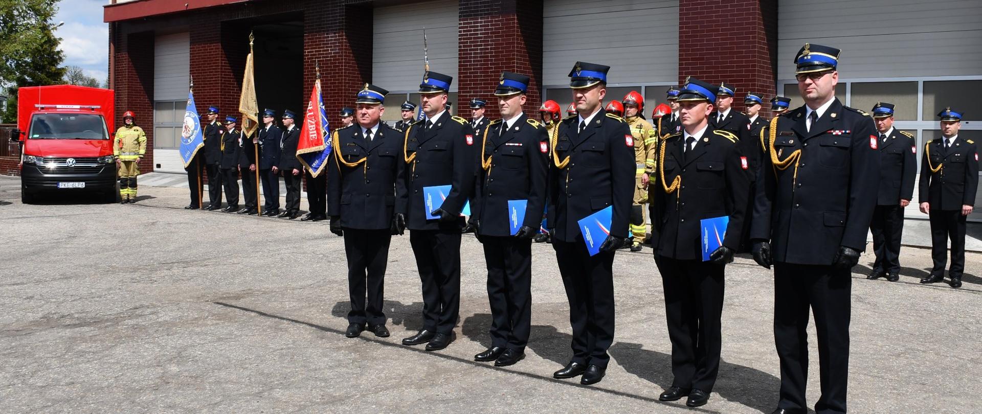 6 strażaków stoi w szeregu przed długim szeregiem innych strażaków, wszyscy w ceimnych mundurach i w rogatywkach, trzymają niebieskie teczki w ręku