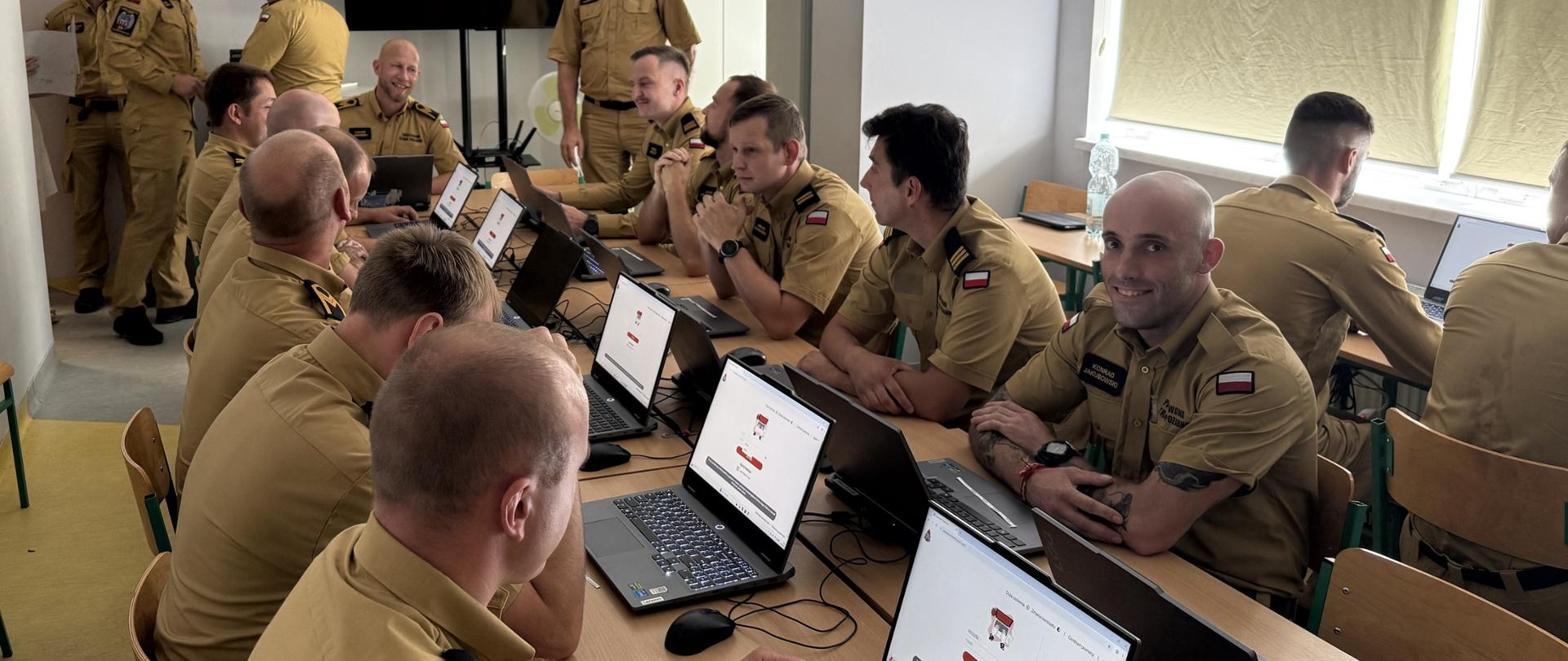 Kilkunastu strażaków w mundurach służbowych, siedzą z obu stron długiego stołu. Przed nimi laptopy - wyposażenie sali komputerowej. W dali kilku strażaków stojących,, ekran tv. 
