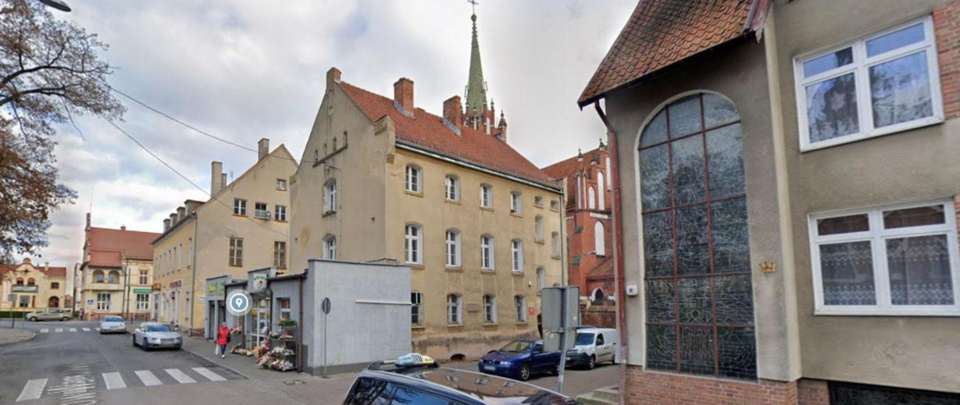 Dwupiętrowy budynek z żółtobladą elewacją i czerwonym dachem. W tle widoczna wieża kościoła.