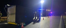 Na zdjęciu widać samochód dostawczy po kolizji w pozycji na boku. Koło pojazdu stoją strażacy a w tle widać samochód pożarniczy oświetlający teren akcji przy użyciu masztu oświetleniowego