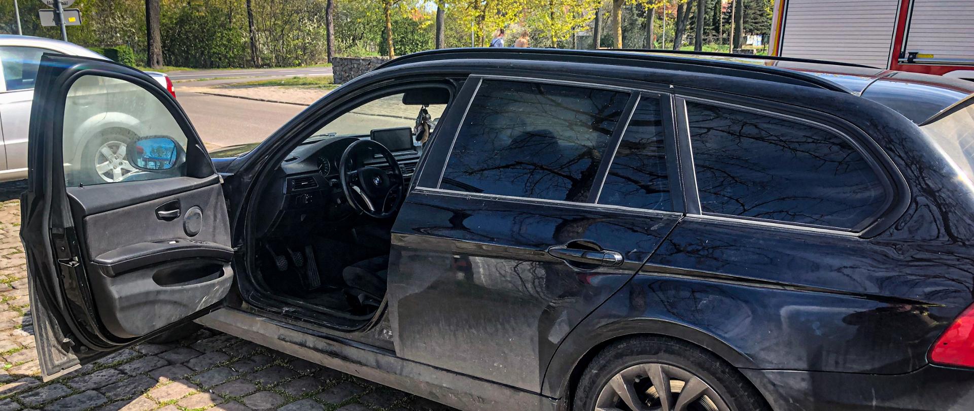 Na fotografii znajduje się czarny samochód osobowy, który ma okopcone szyby oraz otwarte drzwi kierowcy. W oddali znajduje się plac zieleni oraz drzewa, a z prawej strony stoi wóz strażacki.