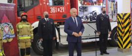 Zdjęcie przedstawia zabierającego głos Wojewodę Lubuskiego na tle stojących w szeregu osób uczestniczących w przekazaniu nowego samochodu ratowniczo-gaśniczego. Od lewej strażak w umundurowaniu specjalnym, Komendant Powiatowy Państwowej Straży Pożarnej w Krośnie Odrzańskim, Komendant Wojewódzki Państwowej Straży Pożarnej w Gorzowie Wlkp. Zdjęcie zrobione zostało w Garażu Jednostki Ratowniczo-Gaśniczej, na tle nowego samochodu.