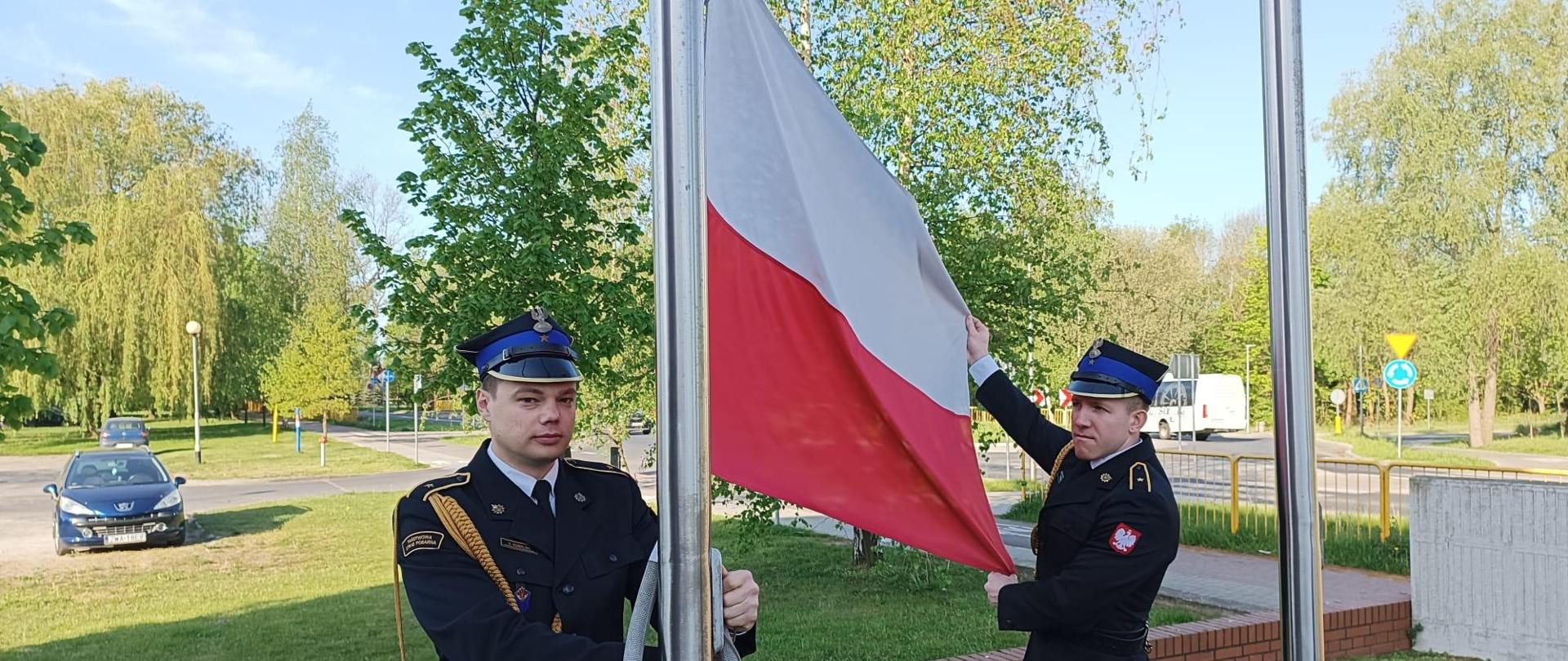 Na zdjęciu widać dwóch strażaków w umundurowaniu reprezentacyjnym wieszających flagę państwową na maszcie znajdującym się przed Komendą Powiatową PSP w Wałczu 