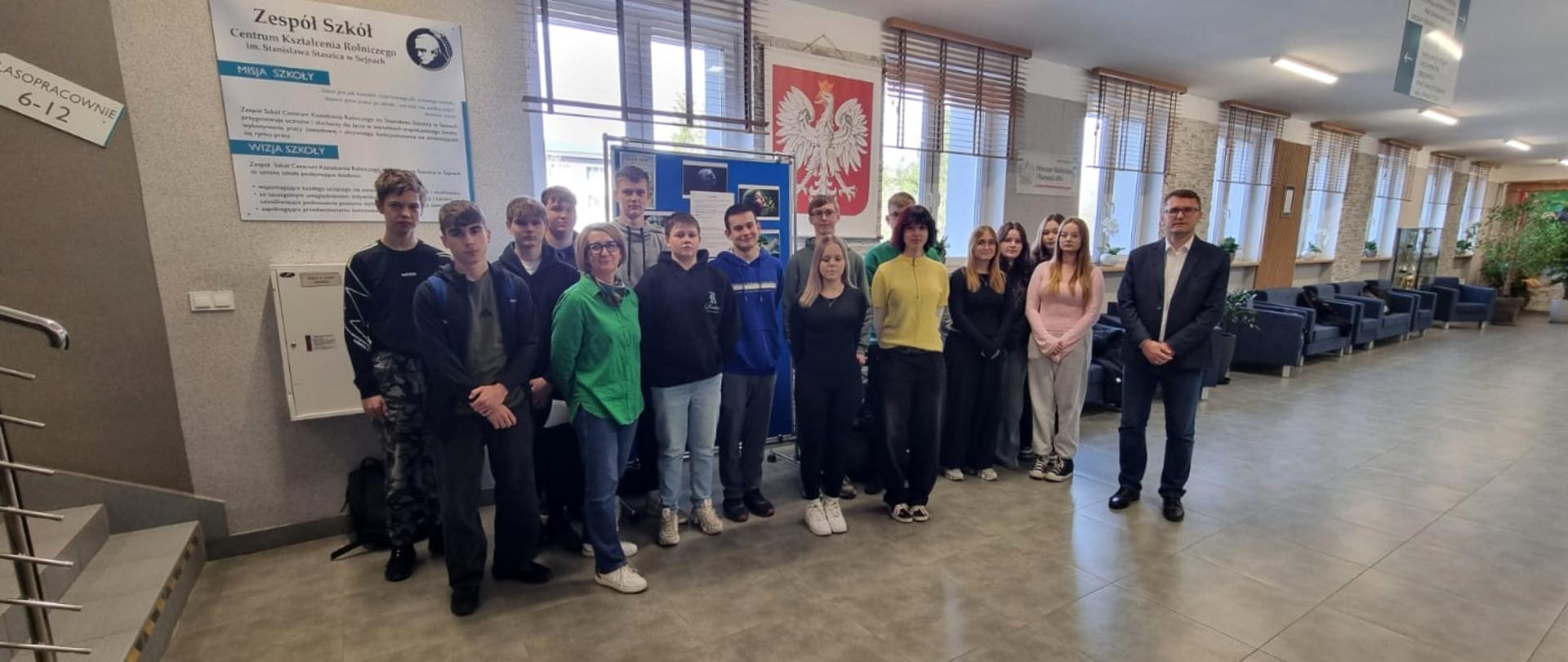 Grupa uczniów oraz dwoje nauczycieli na szkolnym korytarzu.
