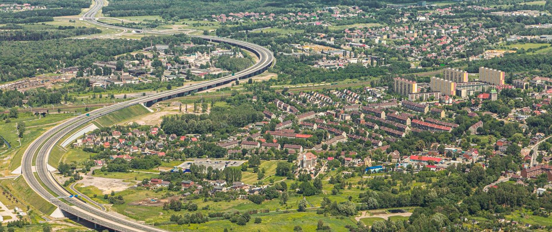 Fotografia lotnicza autostrady A1. Ślad autostrady prowadzony w łukach poziomych, aż po horyzont, w kierunkach naprzemiennych, Autostrada w znacznej części prowadzona długimi obiektami inżynierskimi. W otoczeniu autostrady liczna zabudowa mieszkaniowa poprzecinana terenami leśnymi, w szczególności w północnej części odcinka.