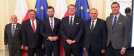 Na zdjęciu znajduje się sześciu elegancko ubranych mężczyzn. Pozują do zdjęcia na tle flag Polski i Unii Europejskiej.