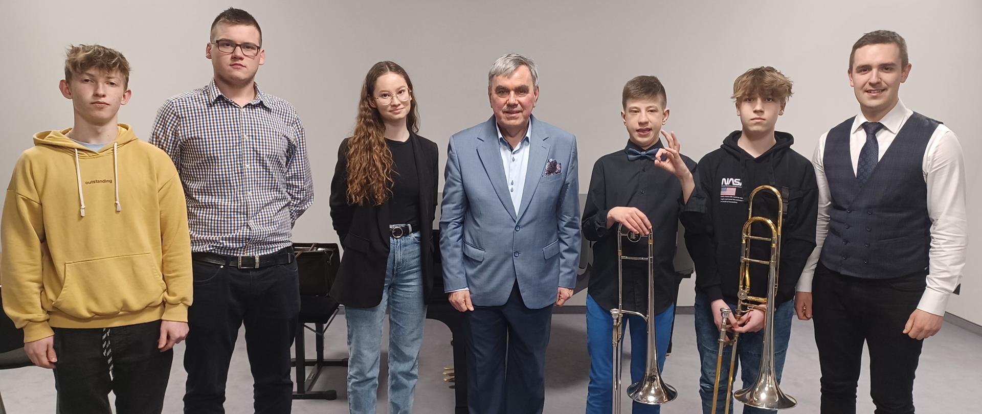 Zdjęcie grupowe. Po środku stoi prowadzący warsztaty puzonowe, po jego prawej i lewej stronie stoją uczniowie biorący udział w warsztatach oraz nauczyciel klasy puzonu. Dwóch uczniów trzyma w swoich dłoniach puzony. Za nimi fortepian oraz ława fortepianowa