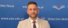 Osoba w jasnym garniturze stoi na tle ścianki promocyjnej Społecznej Akademii Nauk i prezentuje otwarte etui z medalem oraz czerwony dokument. W tle widoczne są logotypy uczelni i adres strony internetowej san.edu.pl.