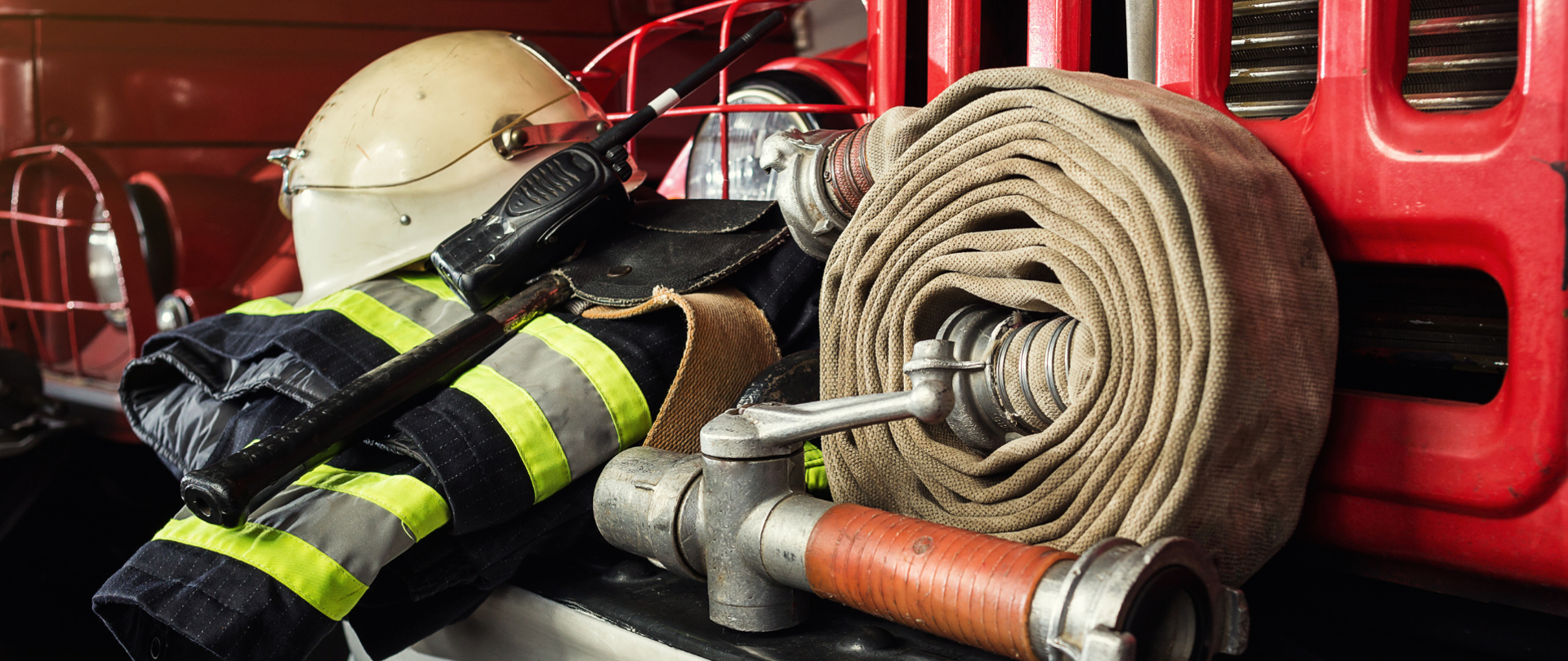 Na zdjęciu znajduje się strój strażacki, kask oraz wyposażenie. W tle widać czerwone wozy strażackie