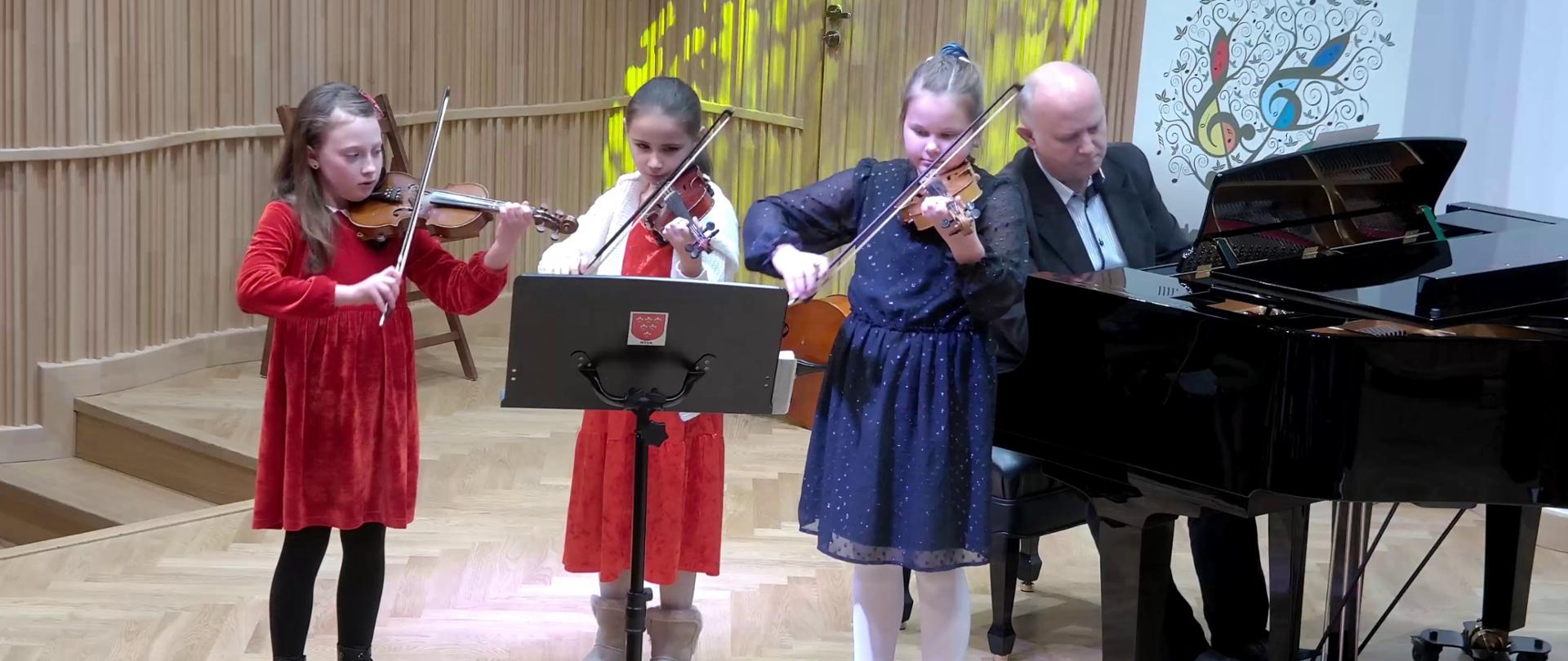 Zdjęcie. Na estradzie występują trzy młode skrzypaczki. Graja z nut ustawionych na pulpicie, który stoi przed nimi. Za nimi na czarnym fortepianie gra ich nauczyciel. W tle po prawej stronie stoi baner PSM w Nysie.