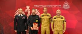 Czterech mężczyzn ubranych w mundury straży pożarnej stoi w szeregu, drugi z prawej trzyma w ręku teczkę o czerwonej okładce z orłem, za nimi widoczny czerwony baner straży pożarnej