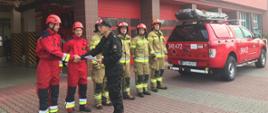 Zdjęcie przedstawia dwóch strażaków ubranych w czerwone kombinezony ratownicze, na głowie czerwone kaski którzy odbierają nagrody od dowódcy JRG1 w Kaliszu. Dowódca obrócony bokiem do kadru, ubrany w mundur strażacki koloru czarnego, czapka strażacka na głowie,, na brzuchu zaciśnięty pas strażacki ściska dłoń jednemu ze strażaków. Obok strażaków odbierających nagrody widzimy czterech strażaków stojących w rzędzie, mających na sobie ubranie specjalistyczne koloru piaskowego, na głowach czerwone chełmy z cyfrą jeden na przodzie. W tle widzimy tył samochód strażacki marki Ford Ranger koloru czerwonego, na klapie bagażnika białe cyfry z nr operacyjnym "341P72",. Na dachu samochodu czarny pokrowiec na sprzęt specjalistyczny. Dalej w tle widzimy otwarte i zamknięte wrota wyjazdowe JRG1 przeszklone i koloru czerwonego.
