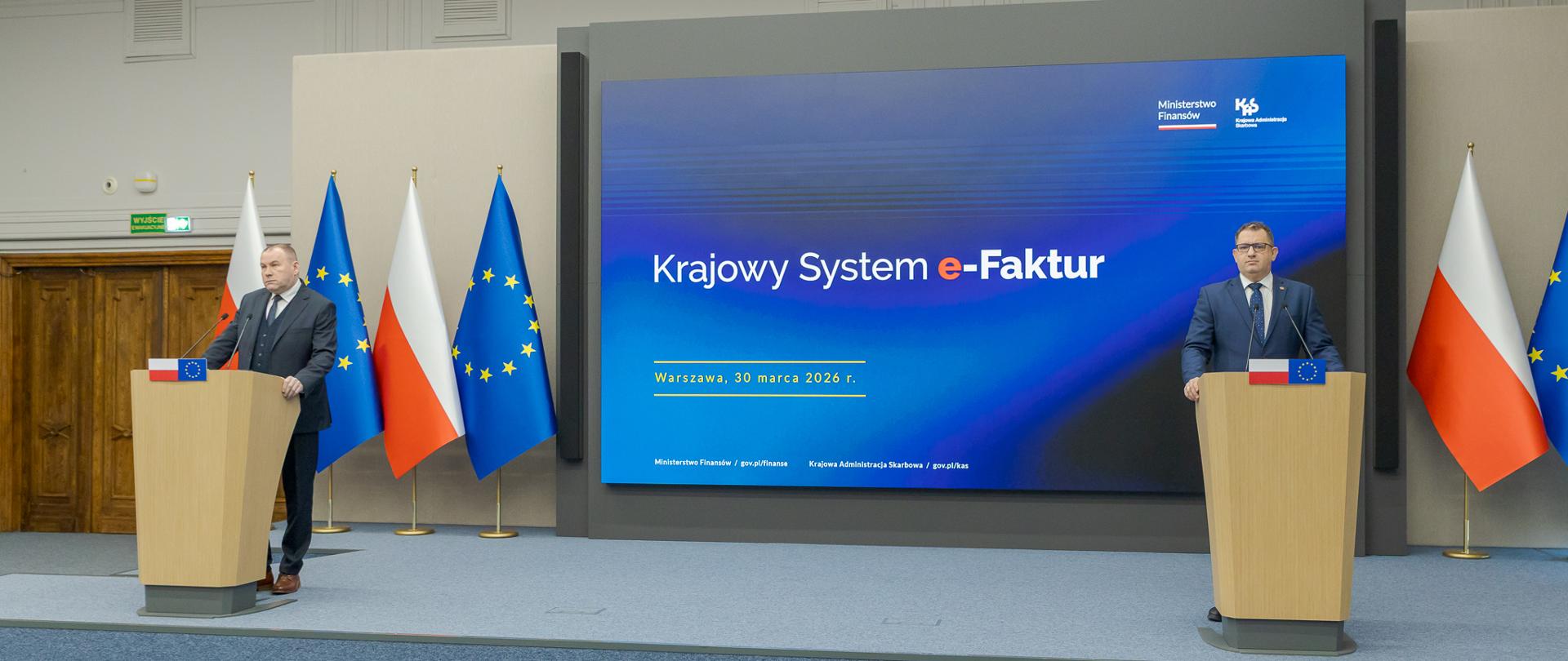 Marcin Łoboda szef Krajowej Administracji Skarbowej oraz Zbigniew Stawicki zastępca szefa KAS podczas konferencji prasowej w Ministerstwie Finansów opowiedzieli o wdrożeniu II etapu Krajowego Systemu e-Faktur przy mównicach przed dużym ekranem z napisem „Krajowy System e‑Faktur”, obok flag Polski i Unii Europejskiej