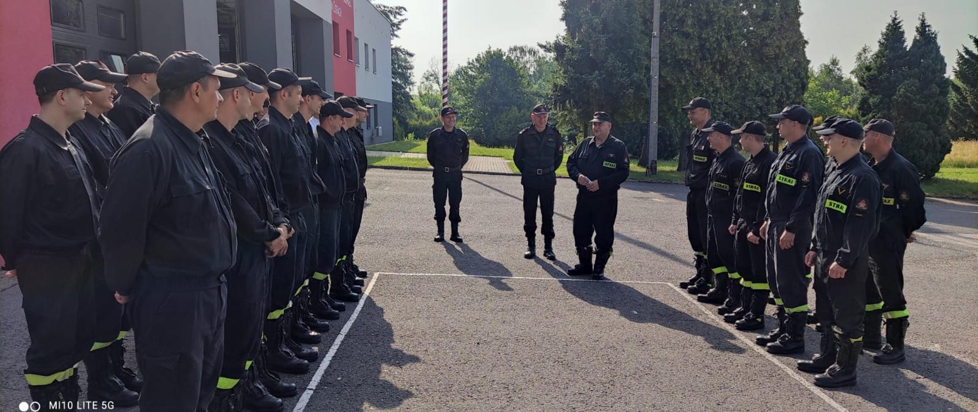 Strażacy stoją w dwóch dwuszeregach twarzami do siebie na placu apelowym. W tle budynek Komendy, drzewa i maszt flagowy.