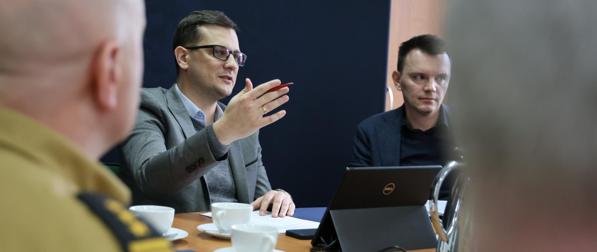 Mężczyzna, gestykuluje ręką, trzymając jednocześnie długopis. Siedzi przy stole, razem z kilkoma osobami. Przed nim tablet. Na pierwszym planie z lewej strony fragment strażaka, widok na pagony.