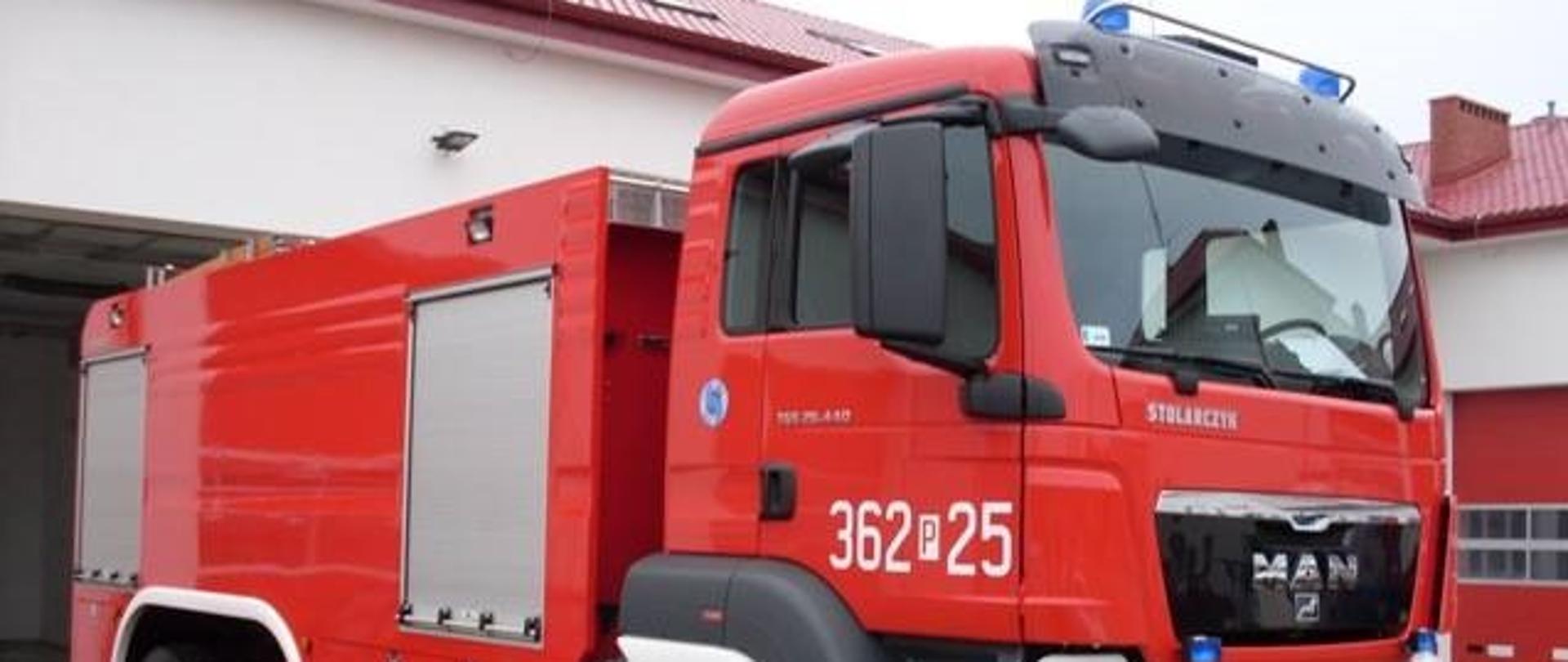 Zdjęcie przedstawia ciężki samochód ratowniczo-gaśniczy z trzyosobową kabiną. W tle zdjęcia widać garaże jednostki ratowniczo-gaśniczej.