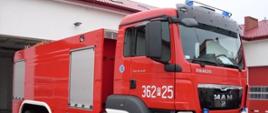 Zdjęcie przedstawia ciężki samochód ratowniczo-gaśniczy z trzyosobową kabiną. W tle zdjęcia widać garaże jednostki ratowniczo-gaśniczej.