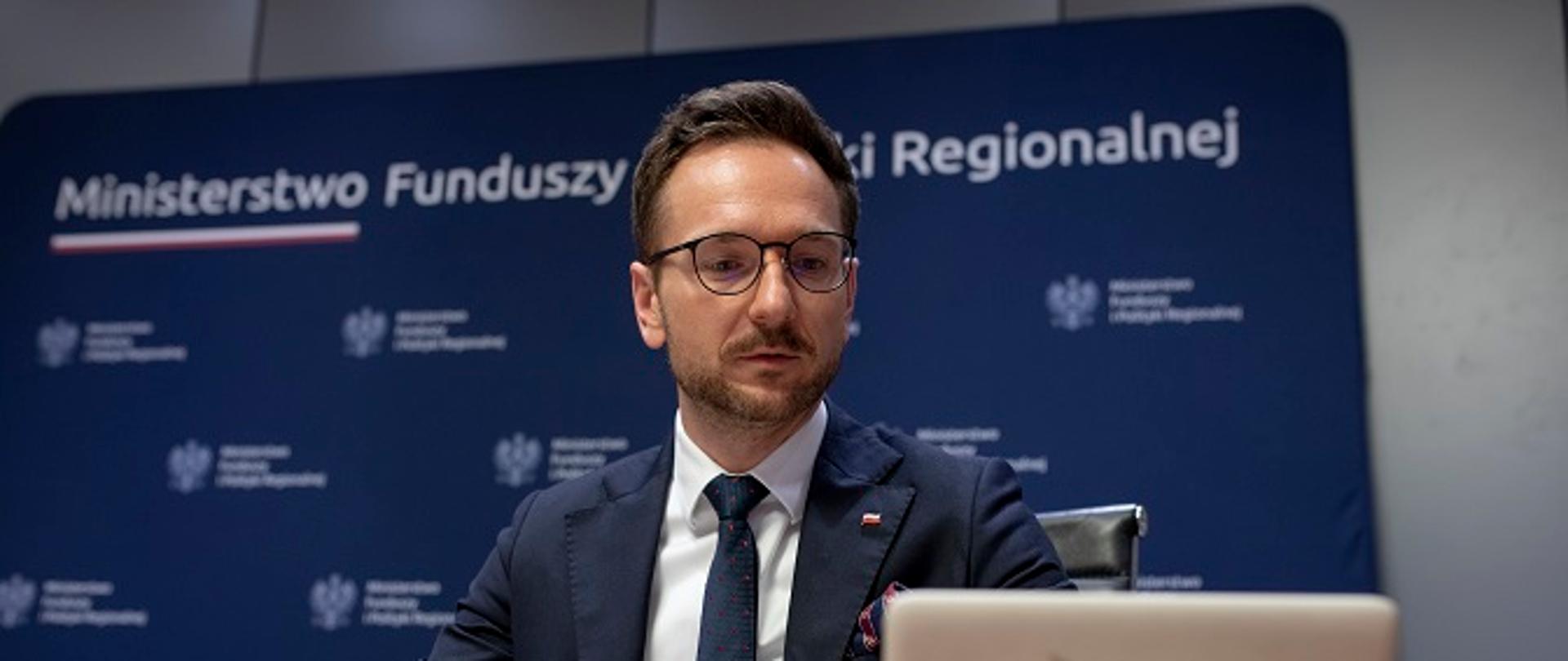 Minister Waldemar Buda siedzi przy stole konferencyjnym, patrzy w tablet. Za plecami ścianka z logo MFiPR.