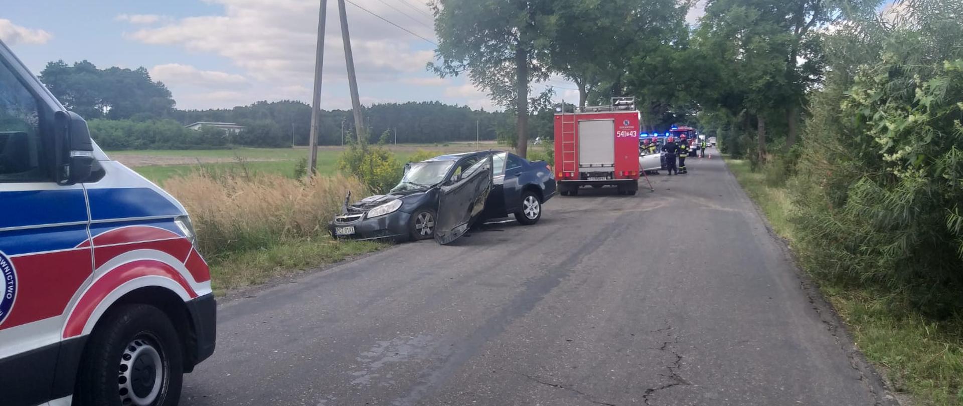 Zdjęcie przedstawia kolizję samochodu osobowego, który uderzył w drzewo. Na zdjęciu widać rozbity pojazd oraz służby ratownicze, które przybyły na miejsce zdarzenia. wrak pojazdu po kolizji. Zdjęcie wykonane w porze dziennej na poboczu drogi.