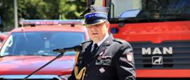 Powiatowe Obchody Dnia Strażaka w Hajnówce - st. bryg. Grzegorz Lisowski podczas swojego przemówienia stoi przy mównicy i zwraca się do zebranych, w tle wozy strażackie