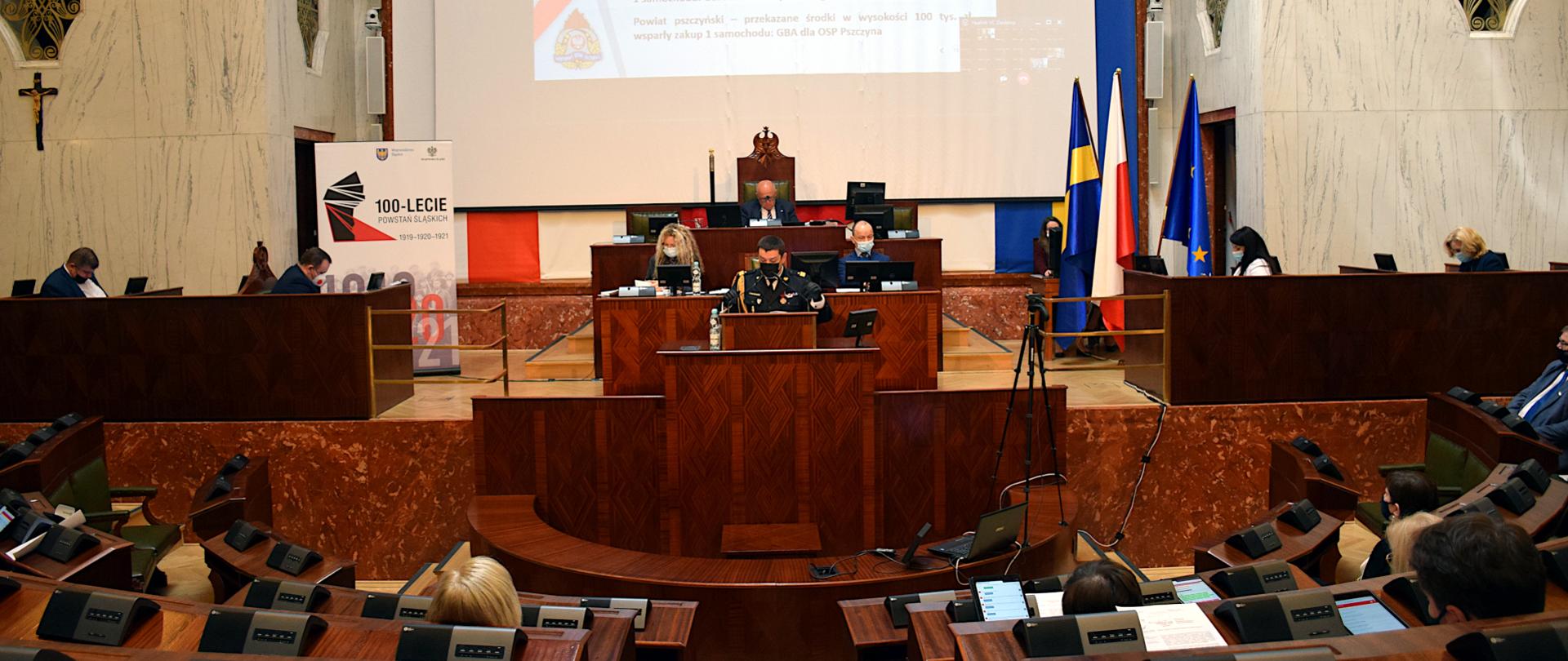 Na zdjęciu widoczna jest sala Sejmu Śląskiego. Na mównicy w mundurze galowym i maseczce ochronnej przemawia Śląski Komendant Wojewódzki PSP. W tle wyświetla się prezentacja. Na sali siedzi między innymi, po lewej stronie w ławkach marszałek i wicemarszałkowie województwa, na wprost przewodniczący sejmiku po prawej członkowie zarządu oraz radni.