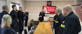 Strażacy i pracownicy cywilni stojący po obu stronach stołu na końcu którego stoi Komendant powiatowy oraz Prezes Zarządu ZOSP RP w Aleksandrowie Kujawskim. Na obrazie wyświetlanym przez TV świąteczna choinka