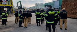 Strażacy Państwowej Straży Pożarnej zapoznają się z charakterystyką zakładu Steico prezentowaną przez przedstawicieli firmy. Osoby znajdują się wewnątrz, obserwują pracę pracowników, niedaleko jest hałda drewna.