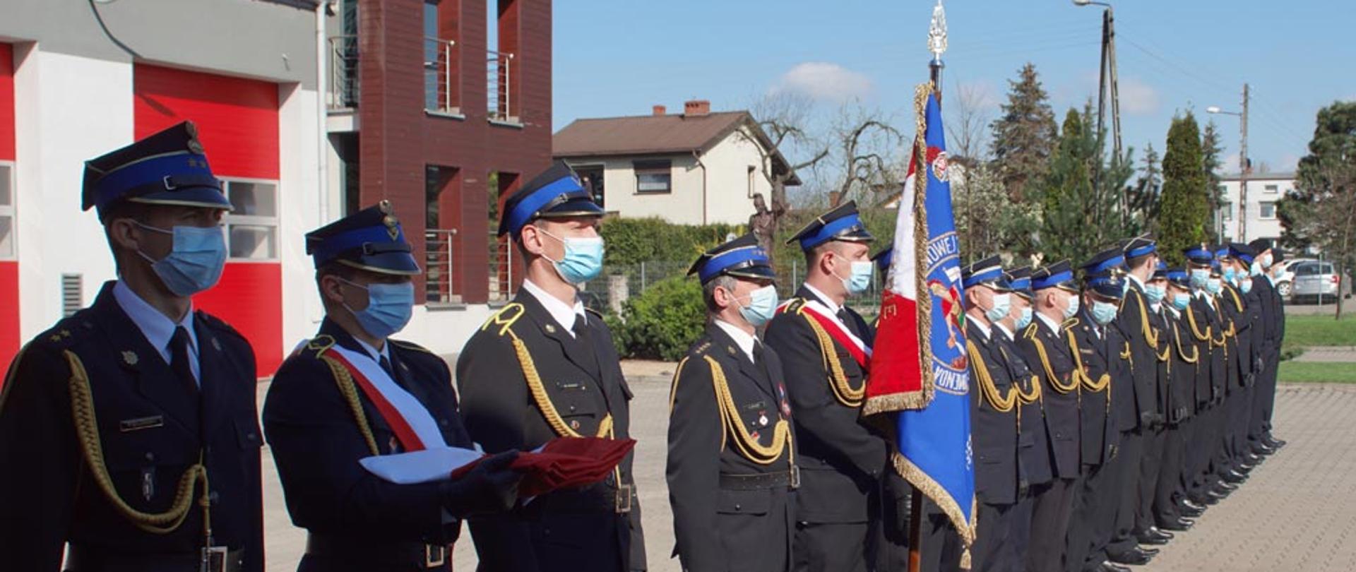 Na placu wewnętrznym komendy stoi pododdział funkcjonariuszy komendy powiatowej Państwowej Straży Pożarnej w Lublińcu. W tle budynek komendy. Na pierwszym planie od lewej stoi poczet flagowy i poczet sztandarowy, obok stoją strażacy w mundurach wyjściowych.
