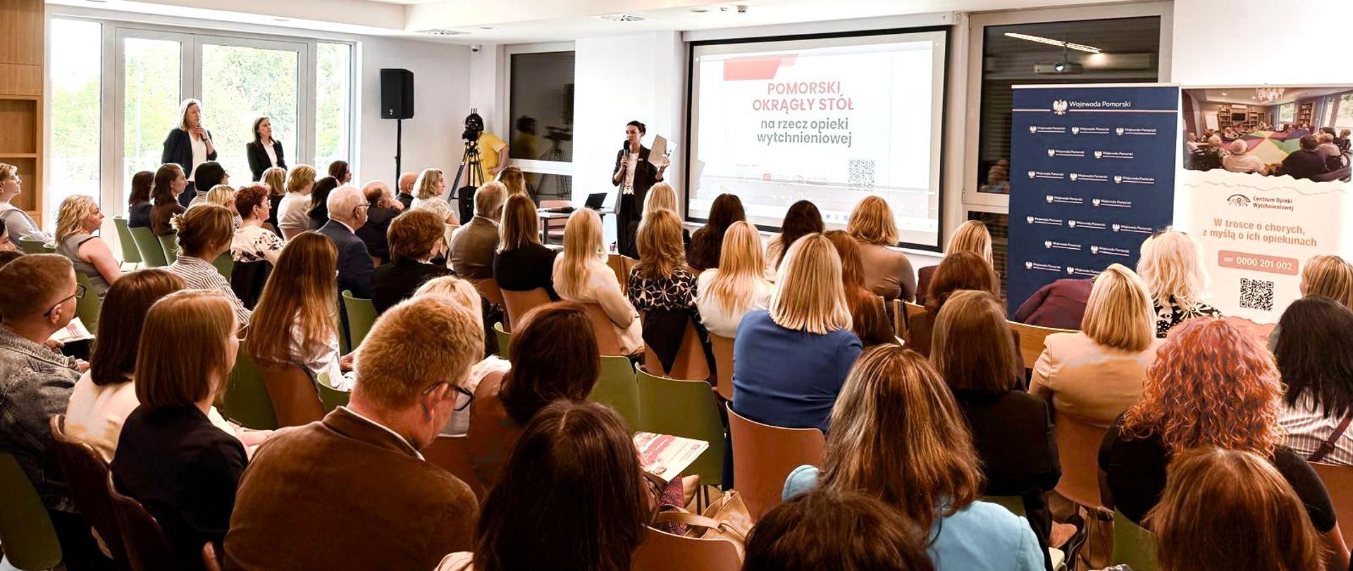Kobieta z mikrofonem prowadzi prezentację przed licznie zgromadzoną publicznością w jasnej sali konferencyjnej. Na wielkim ekranie za prelegentką widnieje napis „Pomorski Okrągły Stół na rzecz opieki wytchnieniowej”. Słuchacze siedzą na krzesłach tyłem do aparatu, skupieni na wystąpieniu.