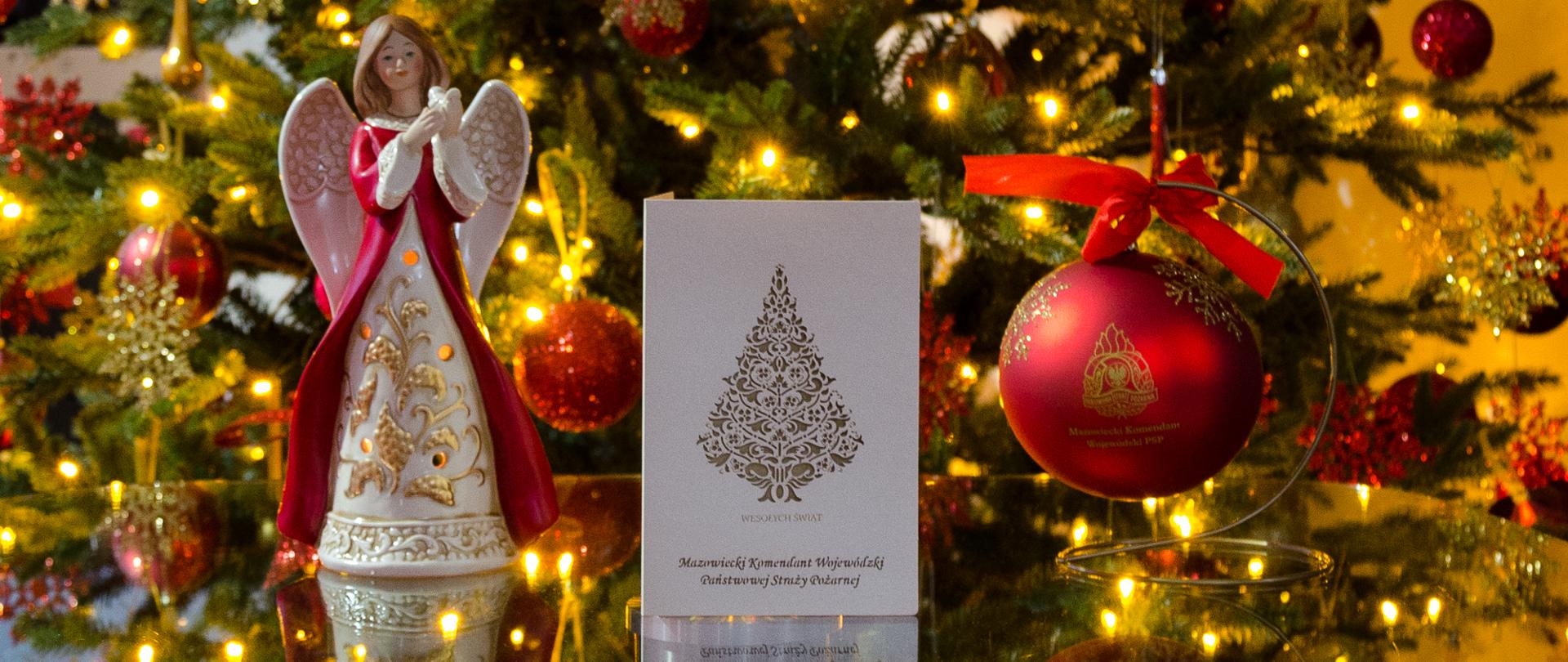Zdjęcie przedstawia beżową kartkę z życzeniami mazowieckiego komendanta wojewódzkiego PSP na święta Bożego Narodzenia 2020. Na kartce wycięta jest choinka. Po prawej stronie zdjęcia stoi czerwona bombka z logotypem PSP na stojaku. Po lewej stronie kartki stoi ceramiczny aniołek w beżowo czerwonej szacie. W tle choinka z lampkami oraz ozdobami.