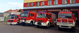 Kolorowa fotografia przedstawia cztery czerwone pojazdy bojowe straży pożarnej przed bramami garażowymi. Pojazdy mają włączone sygnały świetlne. W tle ściana frontowa budynku Komendy Powiatowej Państwowej Straży Pożarnej w Brzozowie. Na ścianie widoczny napis Państwowa Straż Pożarna tel. 998.