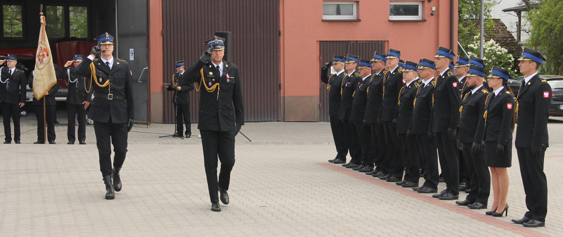 Zdjęcie przedstawia uroczyste obchody Dnia Strażaka z udziałem funkcjonariuszy Państwowej i Ochotniczej Straży Pożarnej podczas apelu na placu przed wewnętrznym komendy. Widoczni są strażacy w mundurach galowych, poczet sztandarowy oraz uczestnicy ceremonii i zaproszeni goście.