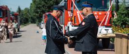Zastępca Komendanta Powiatowego PSP w mundurze wyjściowym przypina medal do munduru galowego strażaka OSP. W tle widać zaparkowane czerwone samochody strażackie.