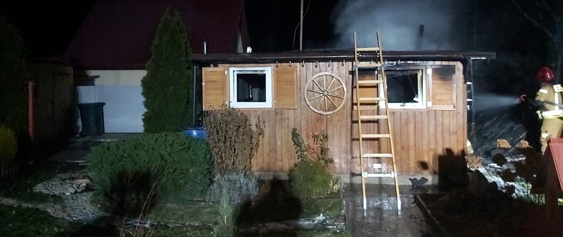 Na zdjęciu domek letniskowy, oparta drabina o jego konstrukcje, po prawej stronie strażak w hełmie koloru czerwonego dogasza pogorzelisko