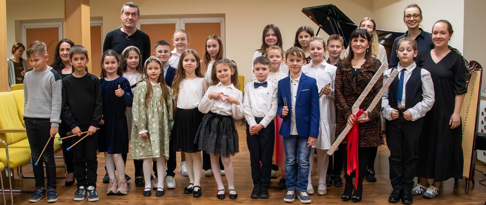 Na zdjęciu grupa kilkunastu osób, dorośli i dzieci, stoją w auli szkoły, za nimi stoi fortepian.