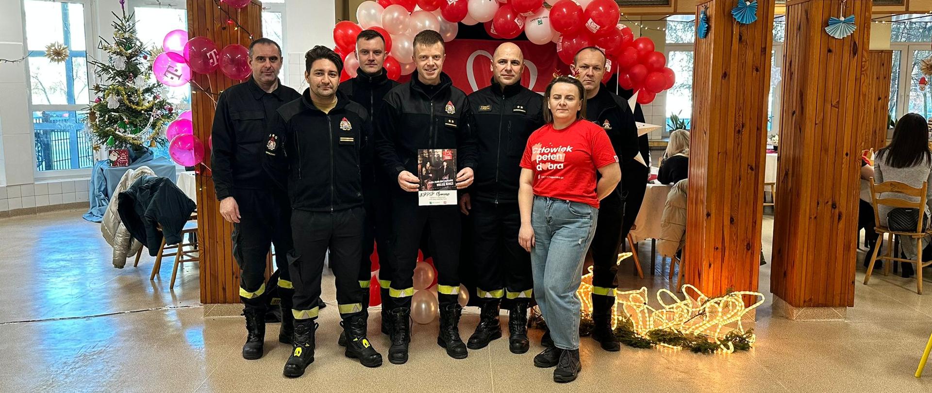 Sześciu strażaków pozuje do zdjęcia w ubraniach koszarowych czarnych, po prawej stronie pozuje również wolontariuszka szlachetnej paczki. Jeden ze strażaków trzyma dyplom szlachetnej paczki. W tle biało czerwone balony nad strażakami i logo szlachtnej paczki. W pomieszczeniu jest również ubrana choinka.