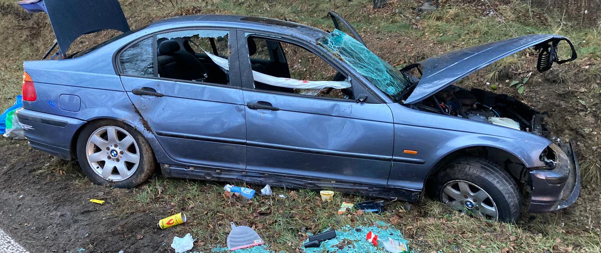Na zdjęciu rozbite auto znajdujące się w rowie. Samochód ma otwartą maskę oraz klapę i zbite szyby