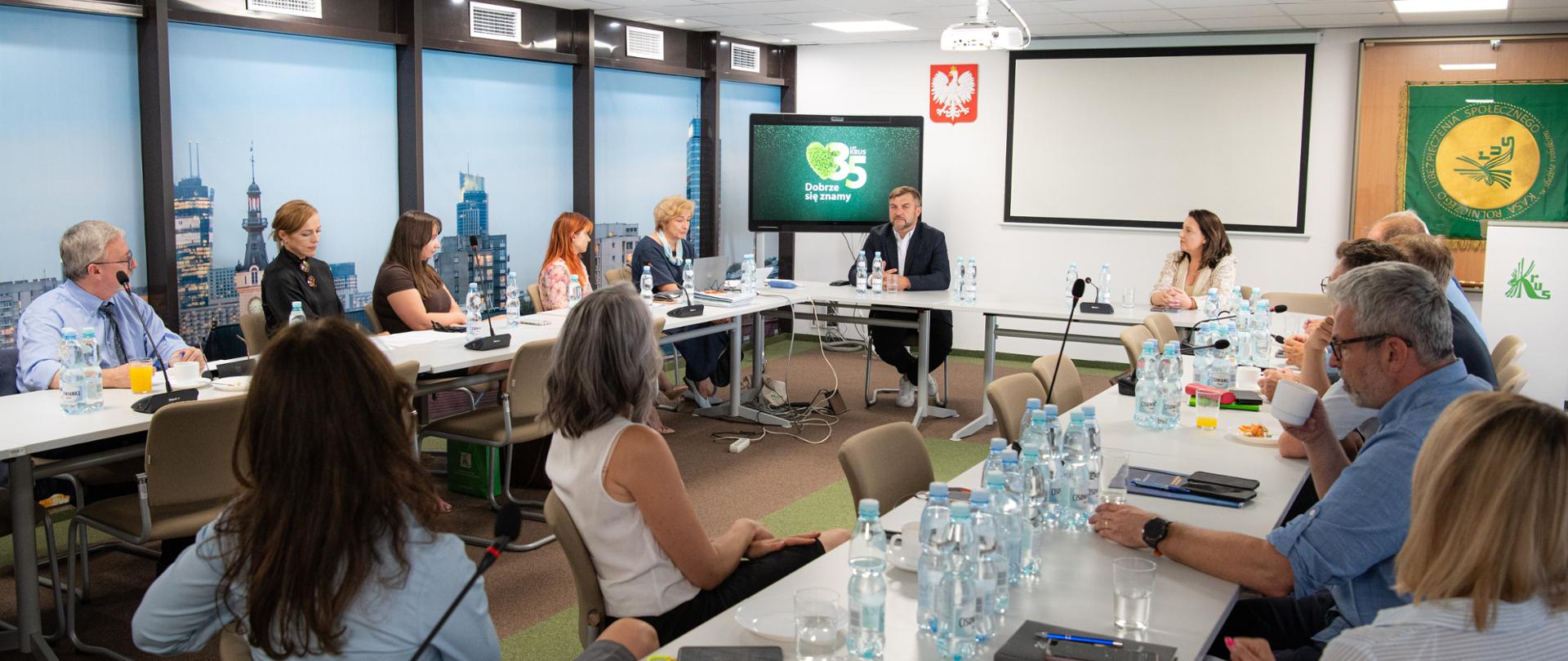 Grupa 14 osób siedzi w sali konferencyjnej przy długich stołach ustawionych w podkowę. W szczycie stołu siedzi mężczyzna w garniturze i białej koszuli obok po prawej kobieta w jasnej marynarce. za nimi od lewej: monitor z logo 35-lecia KRUS, tablica i sztandar KRUS po prawej. 