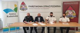 4 mężczyzn siedzących za stołem, podpisuje kartki z porozumieniem. Trzech z nich jest ubranych w mundury, jeden z nich w ciemnym garniturze. Za nimi plansze z napisami "Państwowa Straż Pożarna z logiem Państwowej Straży Pożarnej oraz "Gmina Radomsko"