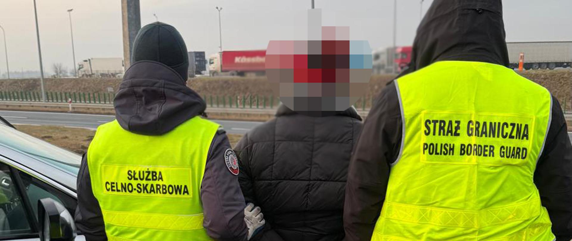 Funkcjonariusze Służby Celno‑Skarbowej i Straży Granicznej stoją przy zatrzymanej osobie.