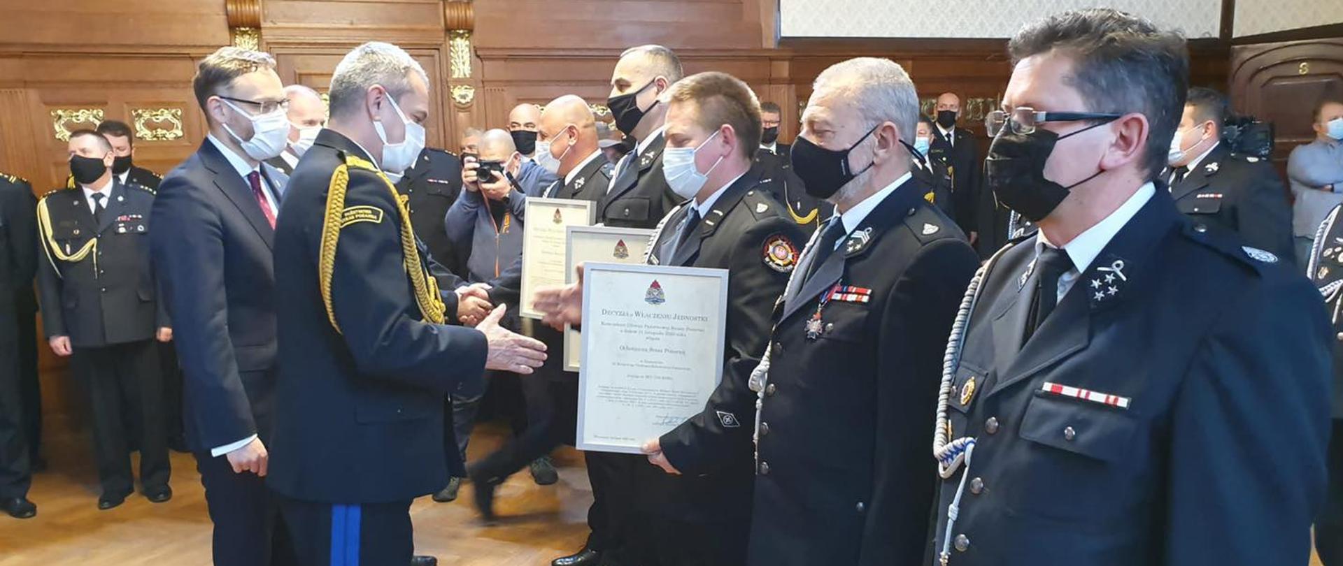 Zastępca Komendanta Głównego PSP wręcza decyzje przedstawicielom OSP, którzy ustawieni są w szeregu,, w tle inni uczestnicy i druhowie
