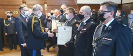 Zastępca Komendanta Głównego PSP wręcza decyzje przedstawicielom OSP, którzy ustawieni są w szeregu,, w tle inni uczestnicy i druhowie