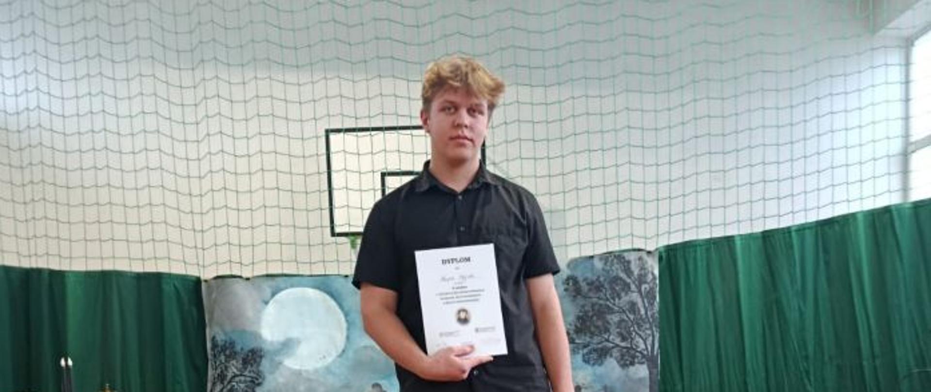 Chłopiec stoi na scenie, trzyma w jednej ręce dyplom, a w drugiej torbę papierową. Z lewej strony fragment stolika z materiałami dekoracyjnymi, obok torby papierowe. W tle siatka zwisająca z sufitu, dekoracja sceny