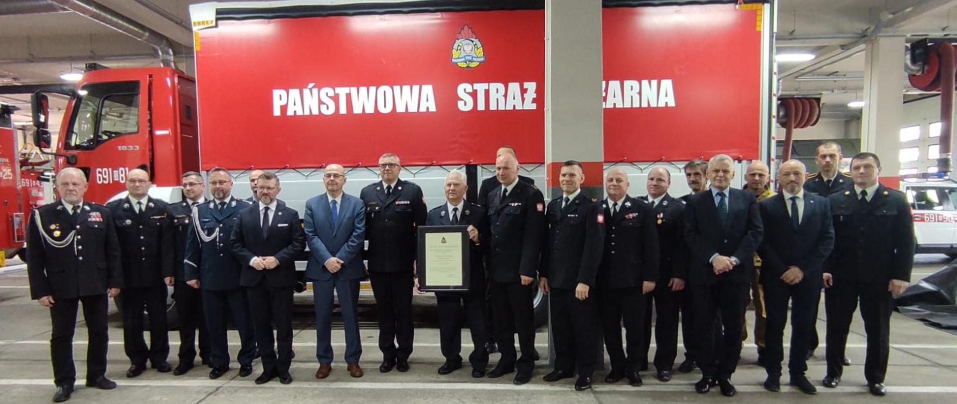 Strażacy i inni uczestnicy narady ustawieni na tle samochodu pożarniczego z logo PSP i napisem Państwowa Straż Pożarna