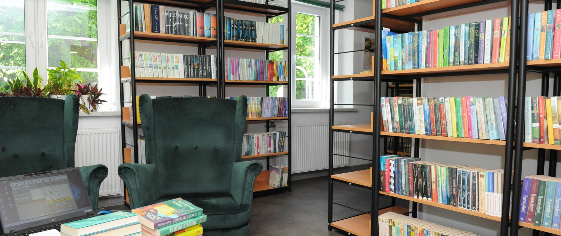Biblioteka szkolna - po prawej stronie stoją dwa regały z książkami i jeden pusty. Na wprost znajdują się dwa okna, a pomiędzy nimi stoją dwa regały z książkami. Na środku ustawione są dwa zielone fotele i okrągły stolik z blatem imitującym zielony granit oraz złotym stelażem. Na stoliku znajduje się laptop oraz dwa stosy po 6 książek.