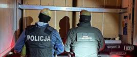 Policjant i funkcjonariusz Służby Celno-Skarbowej stoją przy otwartej naczepie samochodu ciężarowego wypełnionej dużymi kartonami ustawionymi na paletach.